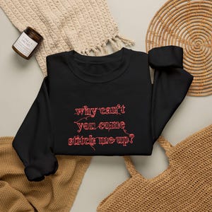 Può includere: Una felpa nera a maniche lunghe con testo ricamato in rosso che recita, why can't you come stitch me up? Le lettere hanno un aspetto strutturato e sfilacciato, e il capo presenta un classico girocollo con polsini e orlo a coste.