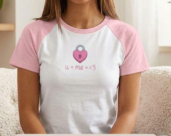 Puede incluir: Una camiseta raglán blanca con mangas y cuello de color rosa claro. El centro de la camiseta presenta un gráfico de un candado rosa en forma de corazón con el texto u + me = <3 impreso debajo en una fuente cursiva rosa claro.