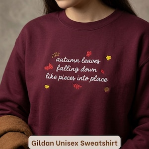 Może przedstawiać: Bordowa bluza z okrągłym dekoltem z białym haftowanym napisem autumn leaves falling down like pieces into place. Wzór jest ozdobiony małymi, kolorowymi haftowanymi jesiennymi liśćmi oraz żołędziem rozmieszczonymi wokół tekstu.