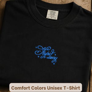 May include: A black crew neck t-shirt featuring blue embroidered cursive text that reads "Stupid sorry" surrounded by small star accents. The shirt has a Comfort Colors brand label at the inner neckline indicating a size large.