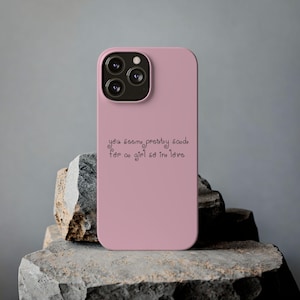 May include: A dusty rose-colored smartphone case featuring a triple-lens camera cutout at the top. The back of the case displays the black text, "you seem pretty sad for a girl so in love," written in a stylized, rounded, and swirling font.