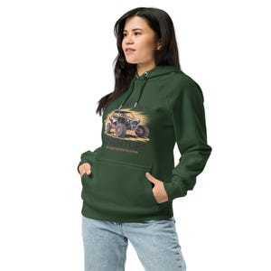 May include: Dark green hoodie with a front graphic of an off-road vehicle, mountains, and the word "ADVENTURE". The hoodie has a drawstring hood and a front pocket. The model is wearing blue jeans.