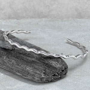 May include: A silver bangle bracelet with a wavy design. The bracelet is open and has a simple, minimalist style.