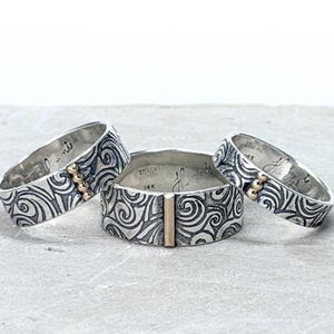 May include: Three sterling silver rings with a swirling pattern and gold accents. Each ring has a different gold accent design. The rings have engraved text. The rings are on a light gray surface.