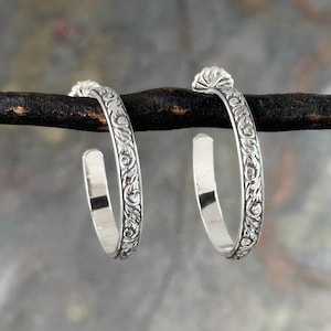 May include: A pair of silver hoop earrings with a floral design. The earrings are about 1 inch in diameter.