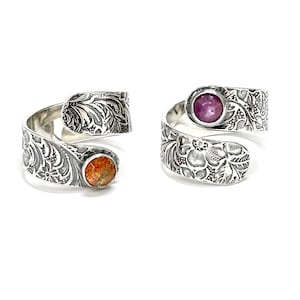 May include: Two silver rings with floral designs. One ring has a purple gemstone and the other has an orange gemstone.