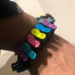 Könnte beinhalten: Ein Armband aus schwarzem Gummi mit bunten Plastikschuh-Anhängern. Die Anhänger haben die Form einer Schuhsohle und sind in den Farben Lila, Blau, Gelb, Rosa und Hellblau.