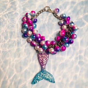Op de afbeelding: Een meerstrengige armband met iriserende roze, blauwe en witte kralen. De armband heeft een zilveren ketting en een bedel in de vorm van een zeemeerminstaart.