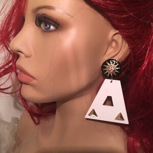 May include: White triangle shaped earrings with a black and gold sun face design.