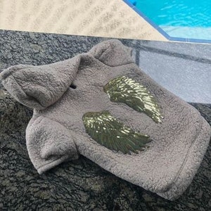 Puede incluir: Un abrigo gris para mascotas con capucha y dos alas bordadas con lentejuelas verdes y doradas. El abrigo está hecho de un material suave y texturizado. El abrigo está diseñado para perros o gatos pequeños.