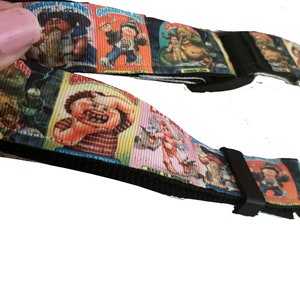 Puede incluir: Una correa de guitarra ajustable negra y blanca con un patrón colorido de personajes de dibujos animados. La correa presenta un patrón repetitivo de imágenes de la serie de tarjetas coleccionables Garbage Pail Kids.