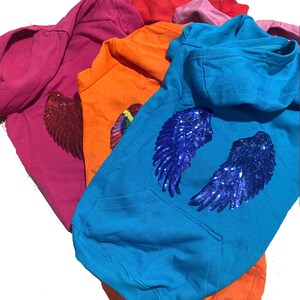May include: A stack of colorful hooded sweatshirts with sequined wings. The sweatshirts are in shades of pink, orange, blue, and red. The wings are blue and silver.