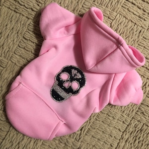 Könnte beinhalten: Rosa Kapuzenpullover für einen kleinen Hund mit einem schwarzen und silbernen Strass-Schädel-Design.