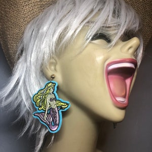 May include: A pair of dangle earrings featuring a mermaid design. The mermaid is embroidered in yellow, purple, and blue on a light blue background. The earrings are made of gold metal.