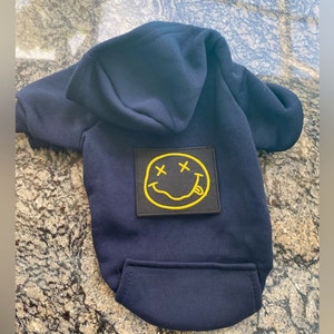 Puede incluir: Una sudadera con capucha azul marino para mascotas con un parche de cara sonriente amarilla con X para los ojos y una lengua que sobresale.