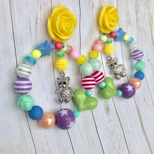 May include: A pair of colorful beaded earrings with a yellow rose charm, a green heart charm, and a silver teddy bear charm. The beads are in various colors, including pink, blue, green, yellow, and purple.