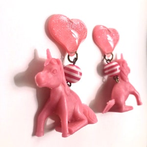 May include: Pink unicorn earrings with a pink heart charm and a pink and white striped bead.
