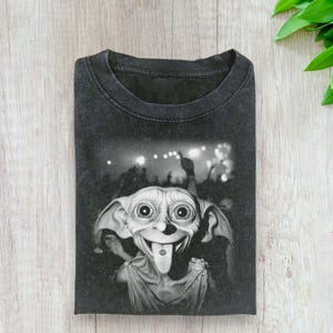 May include: Dark gray t-shirt featuring a black and white graphic of a smiling, cartoon-like creature with a long tongue and a crowd in the background. The shirt is folded and has a vintage, worn look.