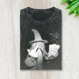 Könnte beinhalten: Schwarzes Grafik-T-Shirt mit einem Zauberer mit langem, weißem Bart, hohem Hut und Pfeife. Der Zauberer hält Geld in der einen Hand und zeigt den Mittelfinger mit der anderen Hand. Das Shirt liegt auf Holz.