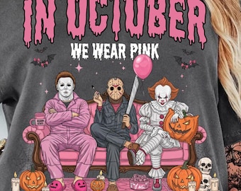 May include: Dark grey t-shirt with the pink text "IN OCTOBER WE WEAR PINK". The graphic shows three horror film characters on a pink sofa, surrounded by pumpkins and bats. The shirt is short-sleeved and casual.