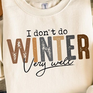 May include: A cream-colored sweatshirt with the text "I don't do winter very well" in brown, gray, and black lettering. The sweatshirt is laid out on a wooden surface with a brown hat, blue jeans, and a small potted plant.