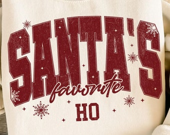 May include: Cream-colored sweatshirt with the text "SANTA'S favorite HO" in burgundy, with snowflake accents. The sweatshirt is paired with blue jeans, a tan hat, and sandals. A small succulent plant is also visible.