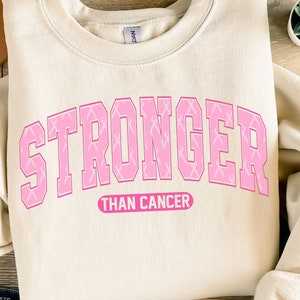 May include: Cream-colored sweatshirt with the text "STRONGER THAN CANCER" in pink, with pink ribbon accents. The sweatshirt is paired with blue jeans, a tan hat, and sandals. A succulent plant is also visible.