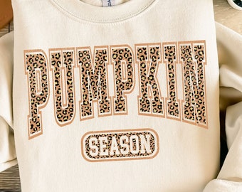 Puede incluir: Sudadera color crema con las palabras "PUMPKIN SEASON" en un diseño de estampado de leopardo. La imagen incluye pantalones cortos de mezclilla azules, un sombrero color canela y sandalias. También se ve una pequeña planta suculenta.