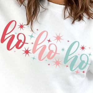 Może przedstawiać: Biała bluza z napisem "ho ho ho" w gradientowym kolorze czerwonym, różowym i turkusowym, z gwiazdkami. Świąteczny design na sezon bożonarodzeniowy.