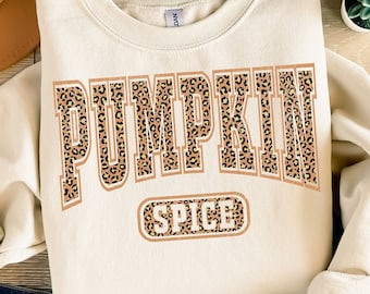May include: A cream-colored sweatshirt with the words "PUMPKIN SPICE" in a leopard print design. The sweatshirt is laid on a wooden surface with a brown hat, blue jeans, and sandals.