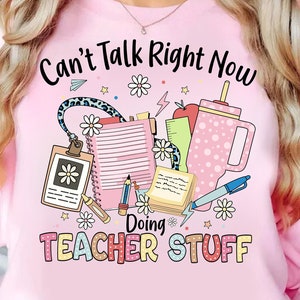 May include: Pink sweatshirt with the text "Can't Talk Right Now Doing Teacher Stuff" and a graphic of school supplies, including a pencil, a ruler, a notebook, a coffee cup, and flowers.