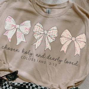 May include: A beige t-shirt featuring three decorative bows with floral patterns. The text "chosen, holy, and dearly loved" is printed below the bows, with "COLOSSIANS 3:12" underneath. A black and white checkered fanny pack, sunglasses, and a scrunchie are also visible.