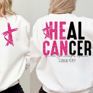 May include: White sweatshirt with a pink cross and ribbon design on the front. The back features the words "HEAL CANCER" in pink glitter and black lettering, with the Bible verse Luke 1:37.