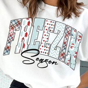 May include: White sweatshirt with a festive design featuring the words "Freezin' Season" in a patchwork style with red, blue, and white plaid, snowflakes, and snowman patterns.