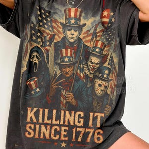 Może przedstawiać: Ciemnoszara koszulka z grafiką przedstawiającą amerykańskie flagi i postacie w patriotycznych strojach. Tekst "KILLING IT SINCE 1776" jest wydrukowany w stylu vintage. Koszulka ma wygląd vintage.