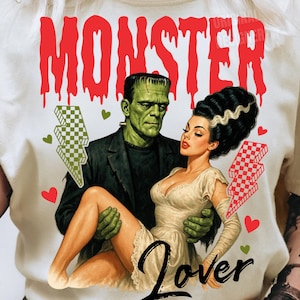 May include: Cream-colored t-shirt featuring a vintage-style illustration of Frankenstein and his bride. The word "MONSTER" is in red dripping letters above the couple. The word "Lover" is below the couple. Green and red lightning bolts are on either side.