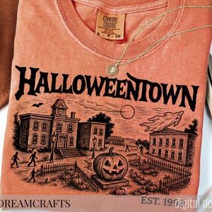 May include: Orange Comfort Colors t-shirt with a black graphic of a Halloween scene, including a jack-o'-lantern, buildings, and the text "Halloweentown." A gold necklace and a brown hat are also visible. The shirt is a digital download.