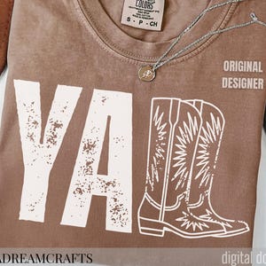 May include: A brown Comfort Colors t-shirt with the word "YA" and a pair of cowboy boots printed in white. The text "ORIGINAL DESIGNER" is printed on the right side. A silver necklace with a pendant is on the shirt.