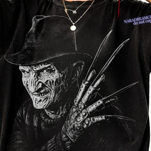 Puede incluir: Camiseta negra con un gr&aacute;fico en blanco y negro de un personaje de pel&iacute;cula de terror con sombrero y guante con cuchillas. El texto "NARADREAMCRAFTS do not copy" es visible en el lado derecho.