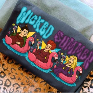 Könnte beinhalten: Dunkelgraues T-Shirt mit einer Grafik von drei Frauen auf rosa Flamingo-Schwimmreifen, die Cocktails halten, mit den Worten "Wicked Summer" in Blau und Lila. Das Design ist vor einem blauen Wasserhintergrund platziert.
