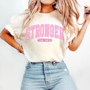 May include: Cream-colored t-shirt with the words "STRONGER THAN CANCER" in pink block letters. The text is in a collegiate style, with a pink ribbon design within the letters. The shirt is paired with light blue jeans.