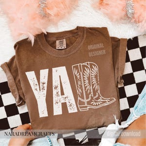 May include: Brown t-shirt with the word "YA" in large white letters and a white cowboy boot graphic. The words "ORIGINAL DESIGNER" are printed on the upper right. The brand name "NARADREAMCRAFTS" is printed on the bottom left.