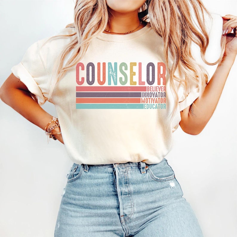 May include: A white t-shirt with the word "COUNSELOR" in colorful retro-style lettering. Below the word are the words "BELIEVER", "INNOVATOR", "MOTIVATOR", and "EDUCATOR" in smaller, colorful lettering.