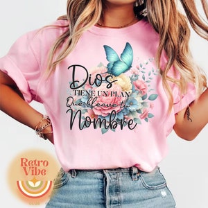 May include: Pink t-shirt with the text "Dios tiene un plan que lleve tu nombre" in black script. A blue butterfly and floral arrangement in shades of pink, yellow, and blue are printed on the front.