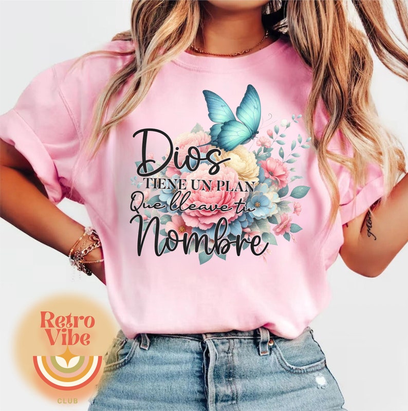 May include: Pink t-shirt with the text "Dios tiene un plan que lleve tu nombre" in black script. A blue butterfly and floral arrangement in shades of pink, yellow, and blue are printed on the front.