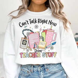 May include: White sweatshirt with the text "Can't Talk Right Now Doing Teacher Stuff" and a graphic of school supplies including a pencil, notebook, ruler, apple, and a cup.