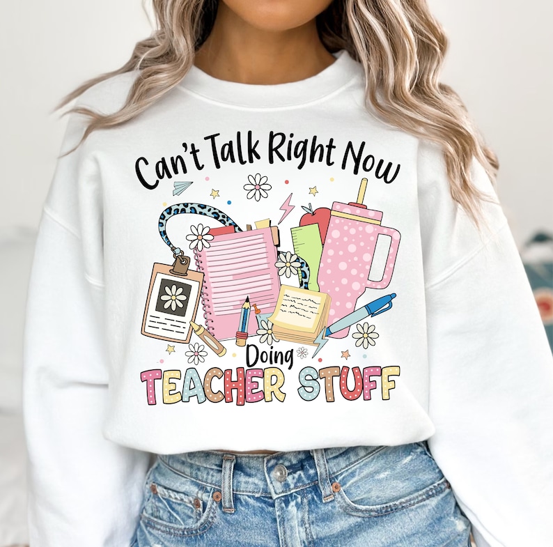 May include: White sweatshirt with the text "Can't Talk Right Now Doing Teacher Stuff" and a graphic of school supplies including a pencil, notebook, ruler, apple, and a cup.