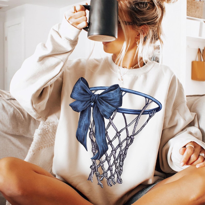 Peut inclure: Sweat-shirt cr&egrave;me avec un motif de panier de basket-ball bleu et un n&oelig;ud. Le motif comprend un n&oelig;ud bleu attach&eacute; au cerceau et au filet. Le sweat-shirt est de style d&eacute;contract&eacute;, adapt&eacute; au quotidien.