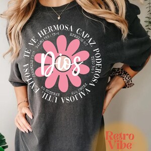 May include: A gray t-shirt with a pink flower graphic and the word "Dios" in the center. The flower has white outlines and the text around the flower is in white. The text reads: "Valiosa Poderosa Capaz Hermosa Util Exitosa Te Ve" with bible verses listed next to each word.