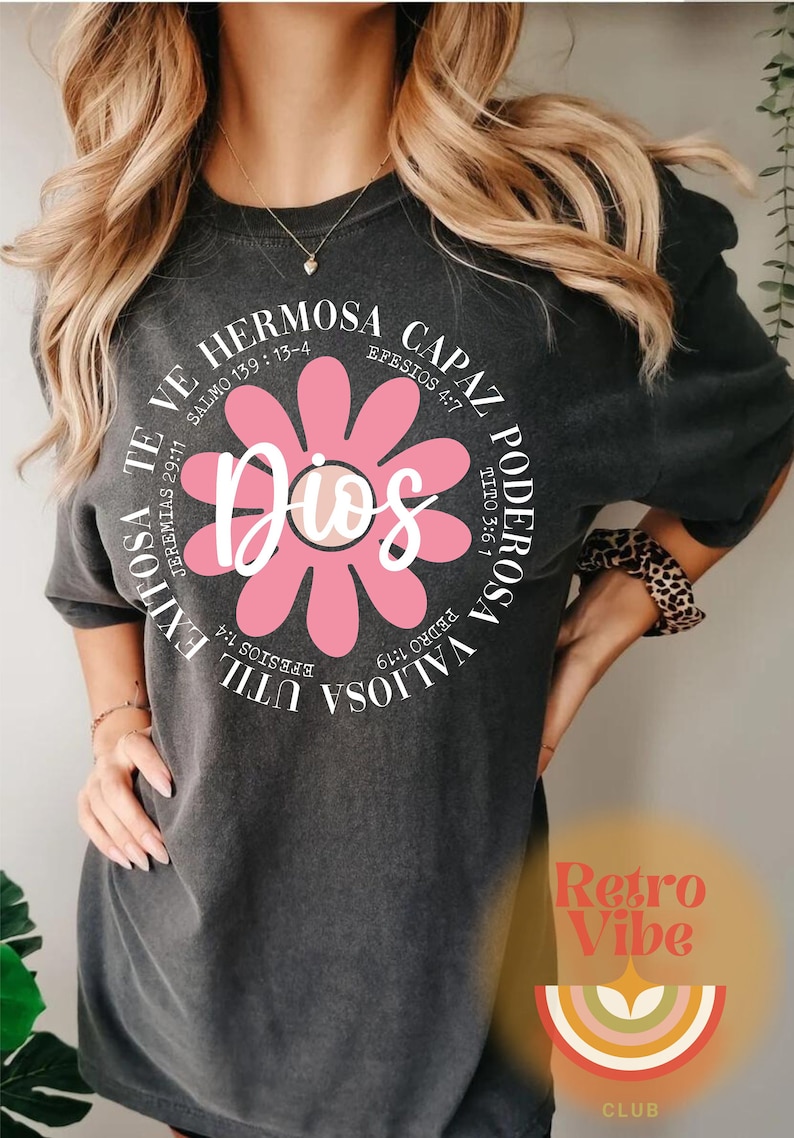 May include: A gray t-shirt with a pink flower graphic and the word "Dios" in the center. The flower has white outlines and the text around the flower is in white. The text reads: "Valiosa Poderosa Capaz Hermosa Util Exitosa Te Ve" with bible verses listed next to each word.